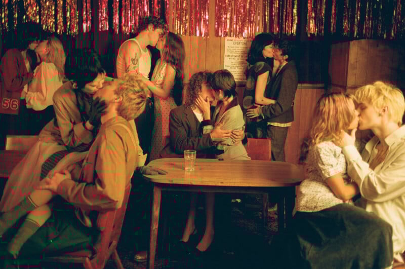 A group of young couples passionately kiss in a dimly lit, festive room with red tinsel hanging on the wall; people sit and stand around a wooden table, creating an intimate and lively atmosphere.