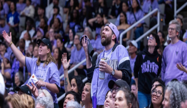 A crowd of fans dressed in purple, some raising hands and cheering, watches an intense moment at a packed indoor sports event.