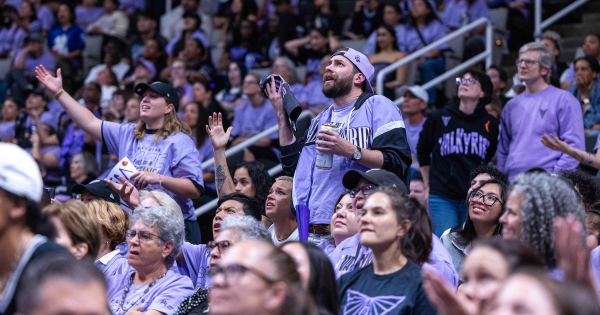 A crowd of fans dressed in purple, some raising hands and cheering, watches an intense moment at a packed indoor sports event.
