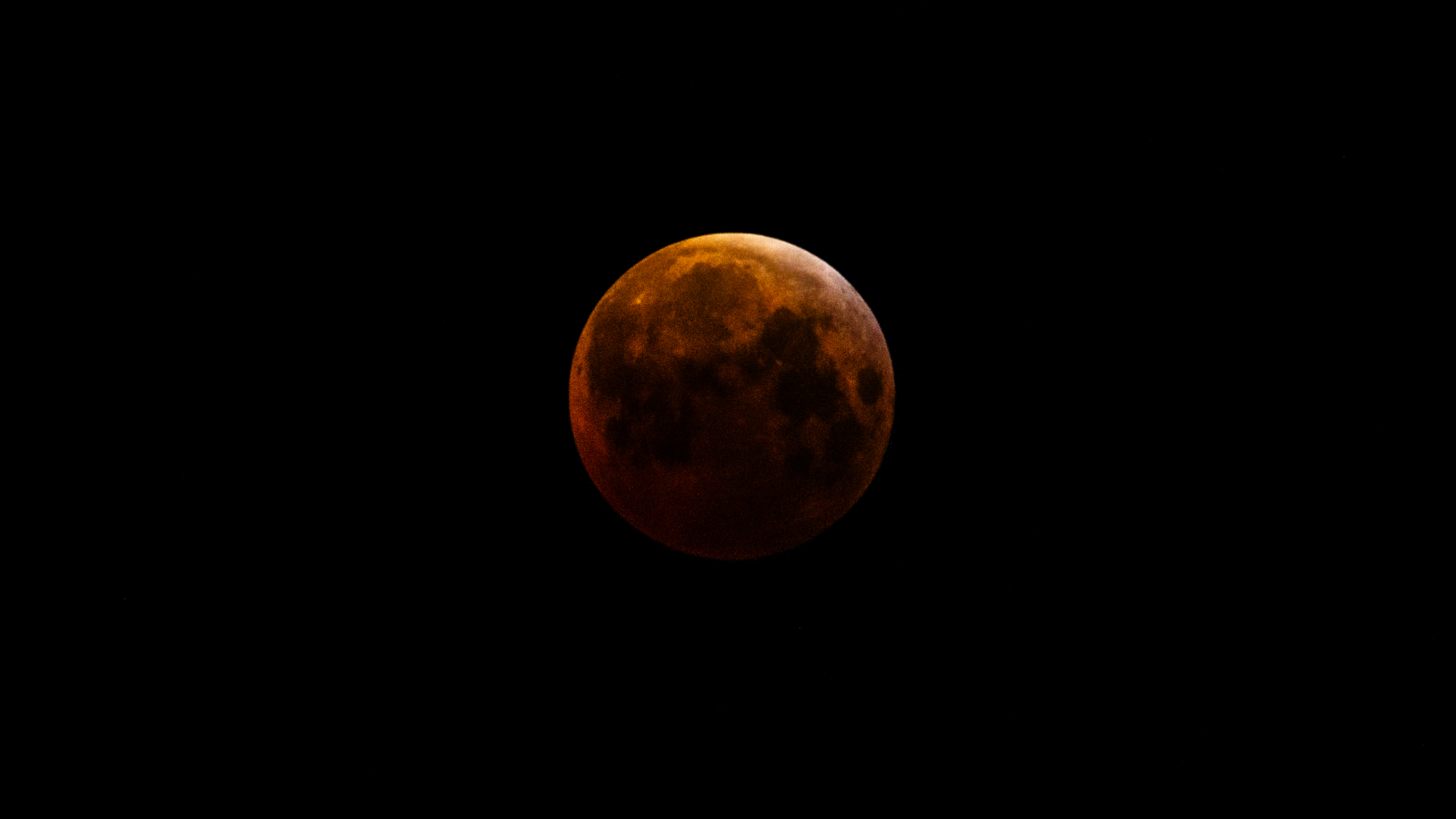 A full moon is pictured during a total lunar eclipse against a black night sky. Earth's shadow has turned the moon a rusty red hue. Lunar maria can also be seen darkening its surface.