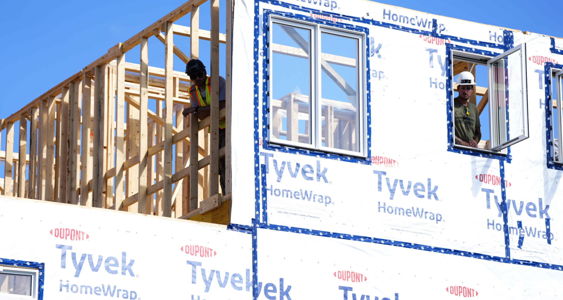 Workers work on a house frame wrapped in plastic against a clear blue sky.