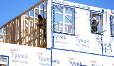 Workers work on a house frame wrapped in plastic against a clear blue sky.