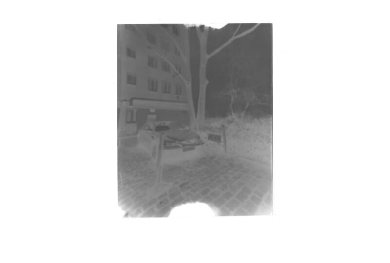 A black and white negative image shows a car parked on a cobblestone street next to a building and trees, with a barrier in front of the car and sidewalk visible in the background.