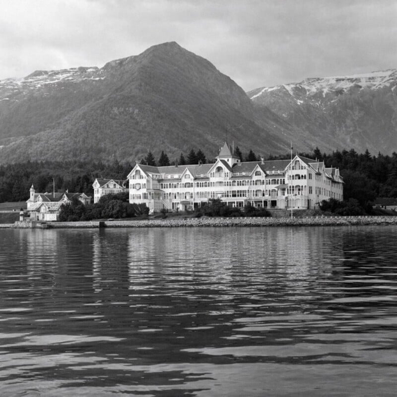A large, ornate building stands on the water’s edge, backed by dense forest and tall, rugged mountains with patches of snow, reflected in the calm lake in the foreground. The image is in black and white.