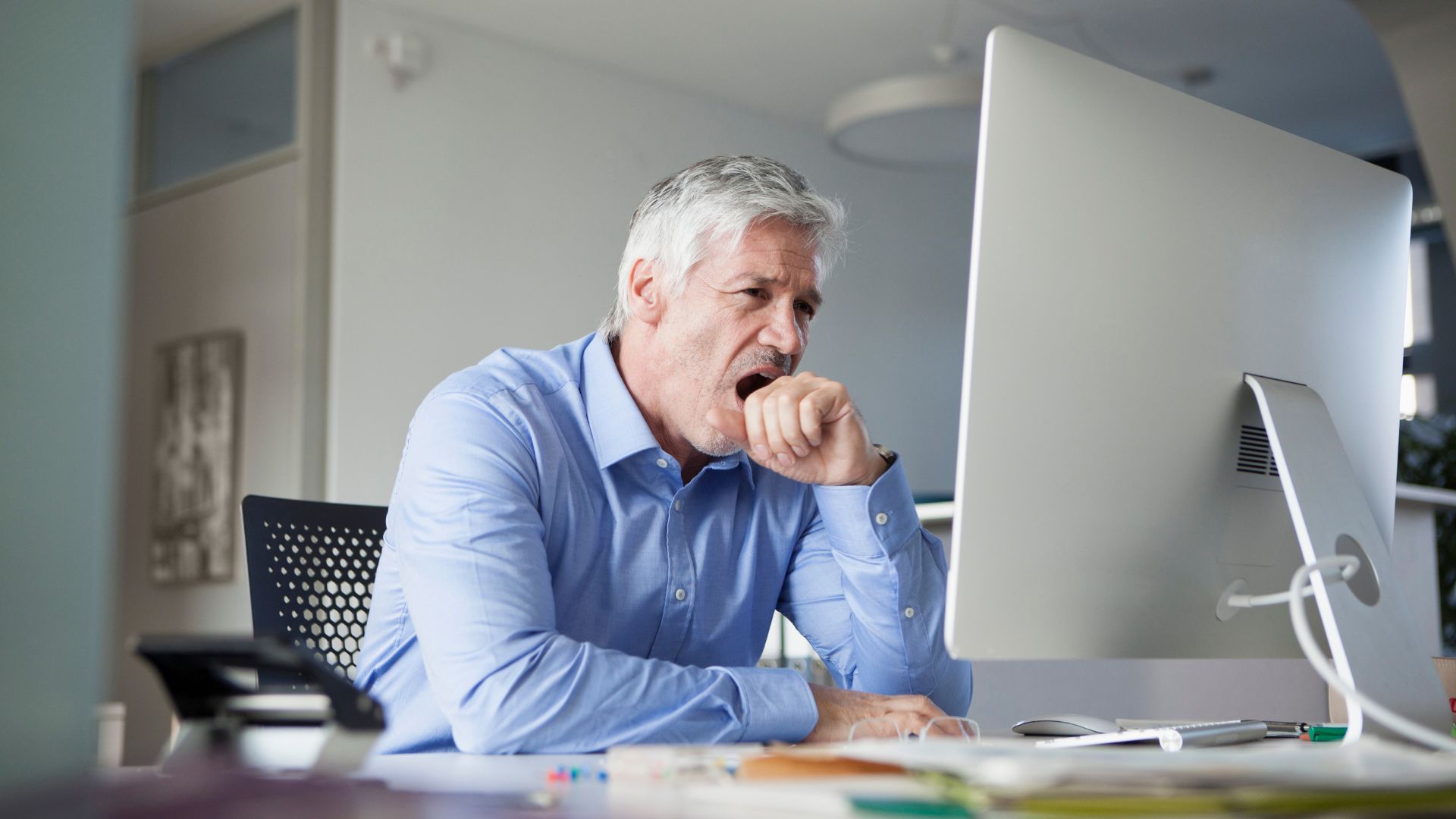 A tired gray-haired man yawns at his desk.