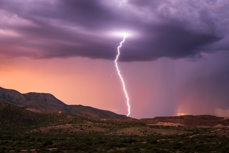 A bright lightning bolt strikes from dark storm clouds above a mountainous landscape with green vegetation at sunset, casting dramatic light over the scene.