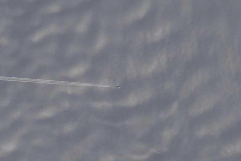 A distant airplane leaves a thin contrail as it flies over a cloudy sky, appearing small against the textured cloud formations below.
