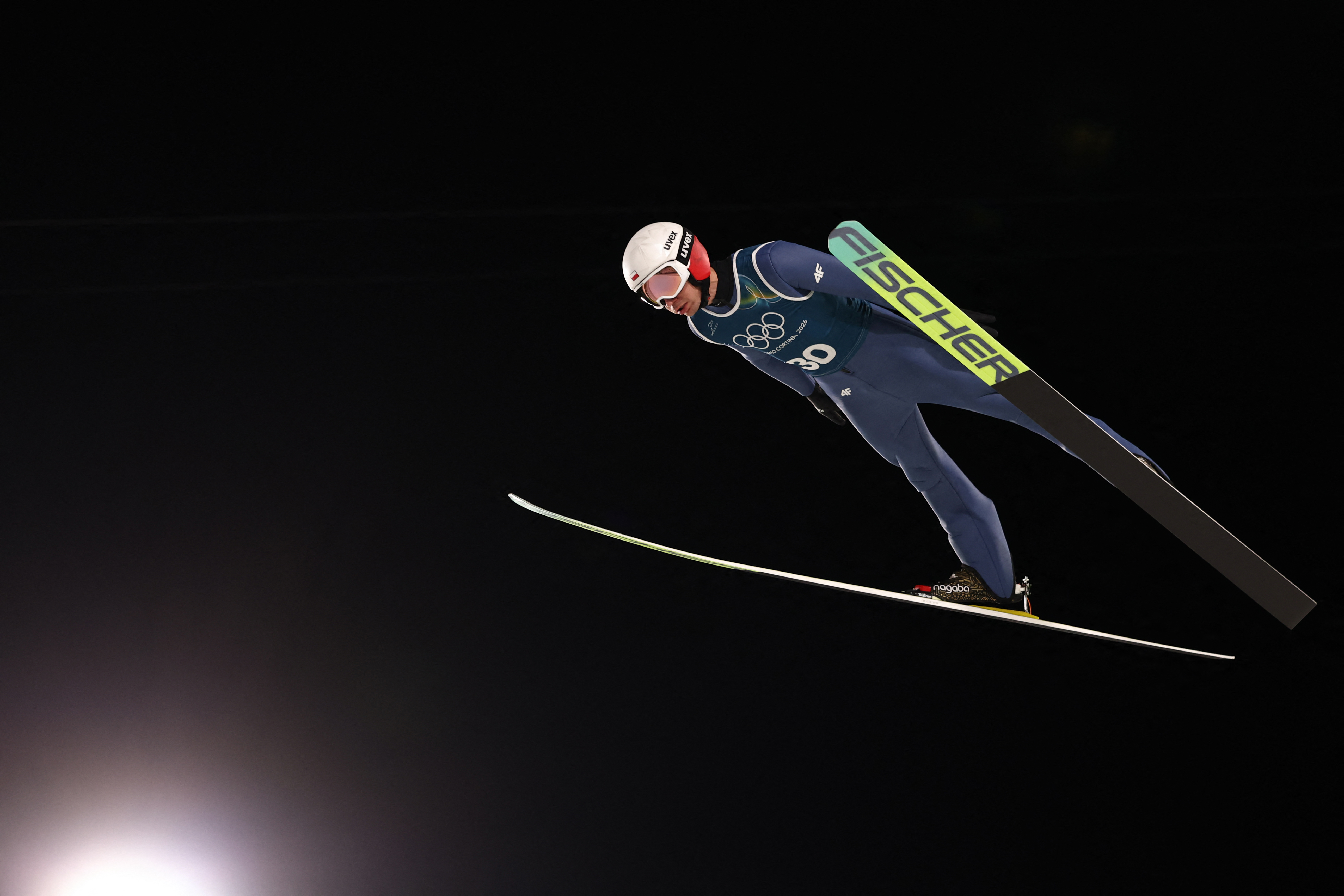 A ski jumper in mid-air against a dark sky wearing a dark blue skinsuit