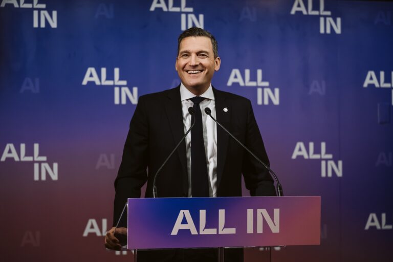Evan Solomon in a black suit and black tie smiles at a podium with an "All In" sign, in front of a purple backdrop with the All In logo repeated numerous times.