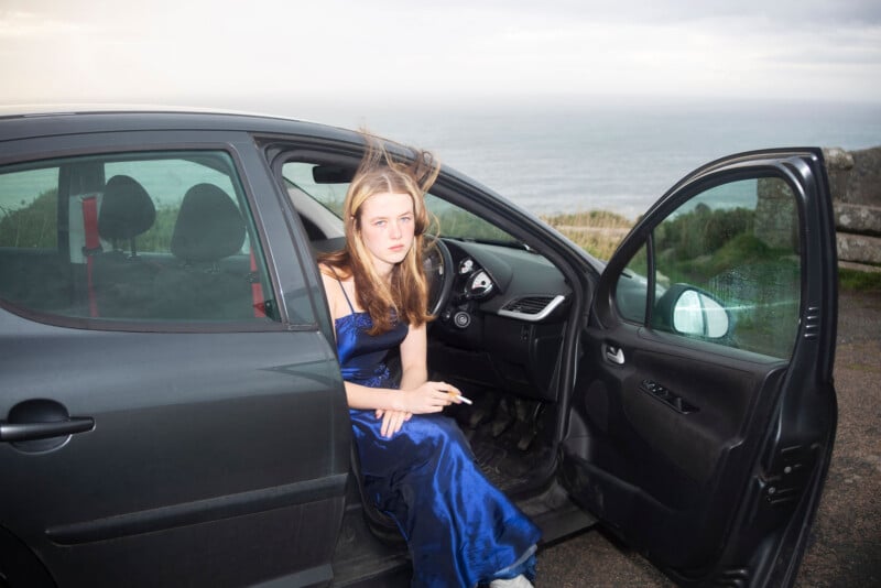 A young woman in a blue dress sits in a car with the door open, holding a cigarette. Her hair is windswept, and the background shows the sea under a cloudy sky.