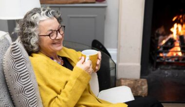 A mature adult sitting by a fireplace in a living room at home. She is wearing a yellow cardigan and spectacles.