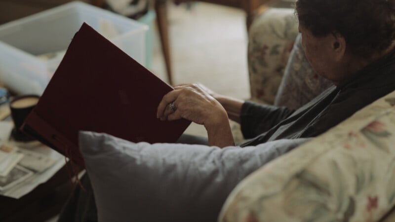 An elderly person sits on a floral couch, holding and looking at a large red photo album. A gray pillow rests nearby, and a plastic storage bin with papers and objects is in the background.