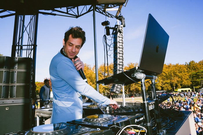 Diplo's Run Club Mark Ronson stood at a DJ deck looking to the side of him. He is wearing a light long sleeved top and holding a microphone.