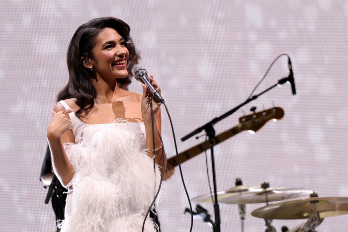 iHeartRadio Hot 99.5's Jingle Ball 2025 Presented By Capital One - Show Olivia Dean wearing a white fluffy dress, smiling as she holda a microphone whil on stage with her band.