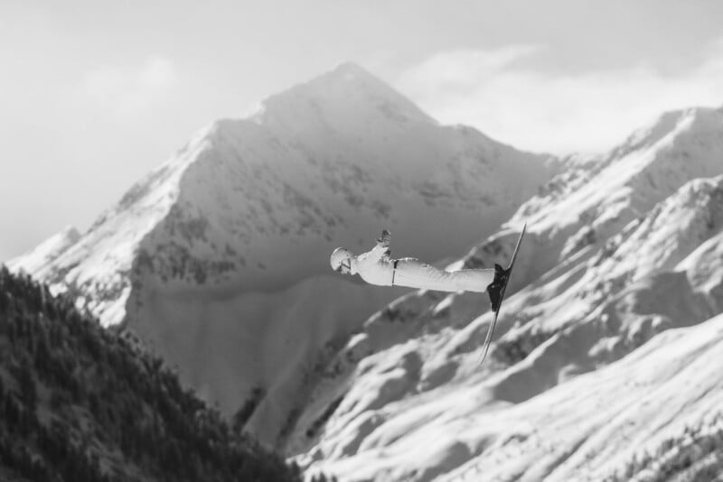 A ski jumper in mid-air performing a jump, with snowy mountains in the background. The image is in black and white, highlighting the dramatic scenery and action.