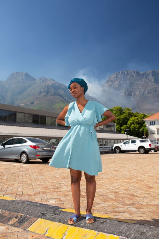 A woman in a light blue dress and sandals stands outdoors on a brick-paved area, with mountains, parked cars, and a modern building in the background under a clear blue sky.