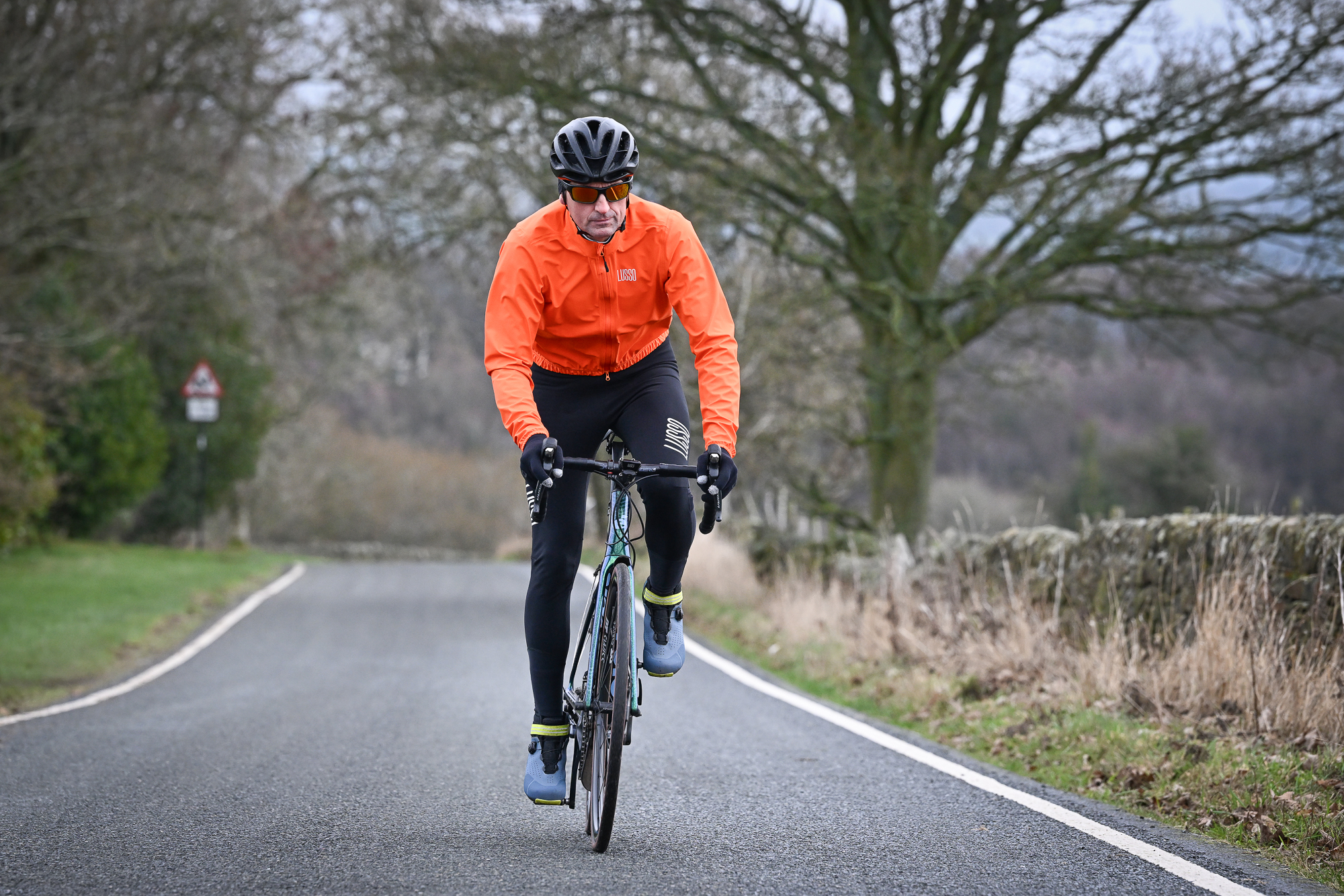 Man wearing black tights, blue shoes and an orange jacket riding a green road bike towards the camera