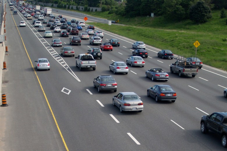 A few cars drive in a high occupancy lane at the left while there are far more cars in the other five lanes at the right. The HOV has white diamond shapes on its pavement to indicate its for vehicles that are carrying more than one person.