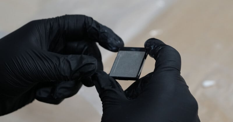 A person wearing black gloves holds a small rectangular object, possibly a microscope slide or electronic component, against a blurred, neutral background.
