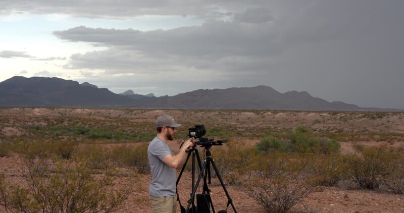 A person in a gray shirt and cap sets up a camera on a tripod in a desert landscape with sparse vegetation, distant mountains, and a cloudy sky.