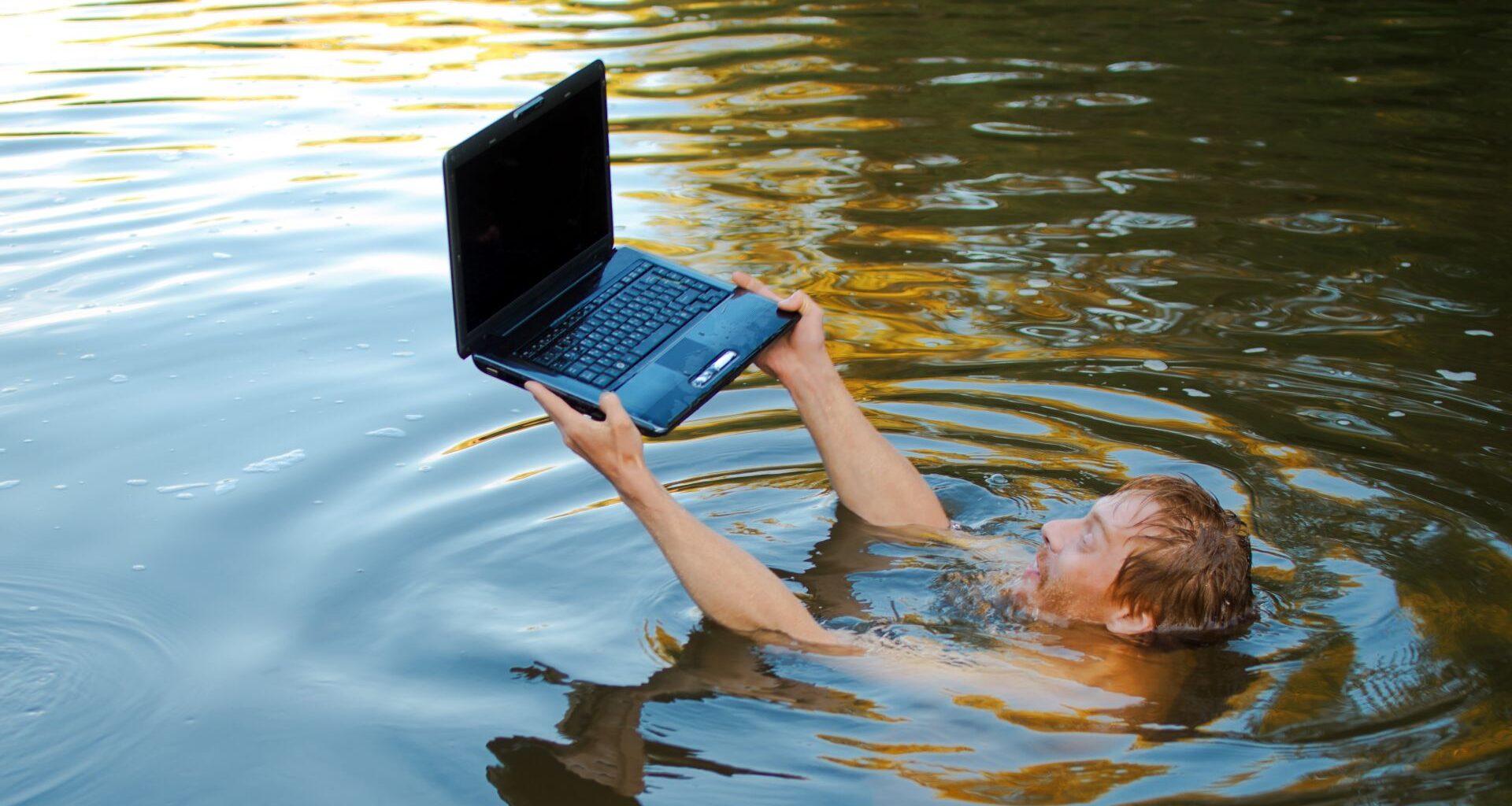 A man submerged in water holding a laptop above the surface.