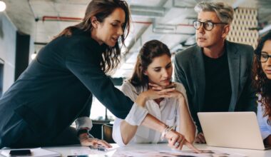 A group of people in business surrounding leaning over a table with laptop and papers on it.