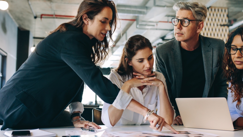 A group of people in business surrounding leaning over a table with laptop and papers on it.