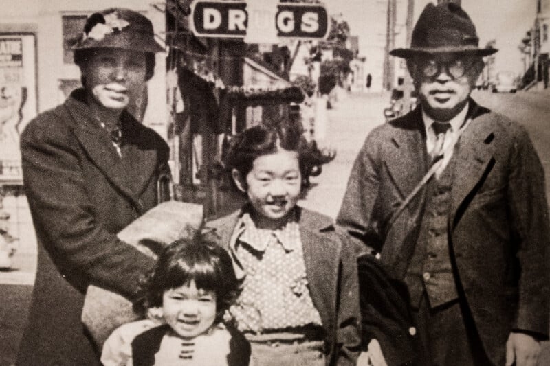 A black-and-white photo of a family of four, two adults and two children, standing together on a sidewalk in front of a building with a "DRUGS" sign. They are all wearing coats and hats, smiling at the camera.