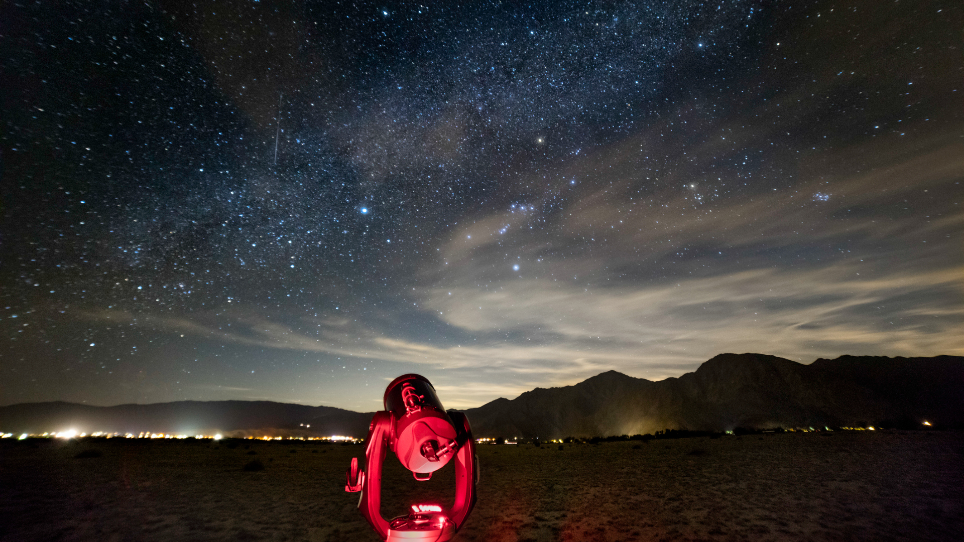 A telescope is illuminated in dim red light in the foreground where a silhouette of mountains is in the background, all under a blue and white streaked starry night sky