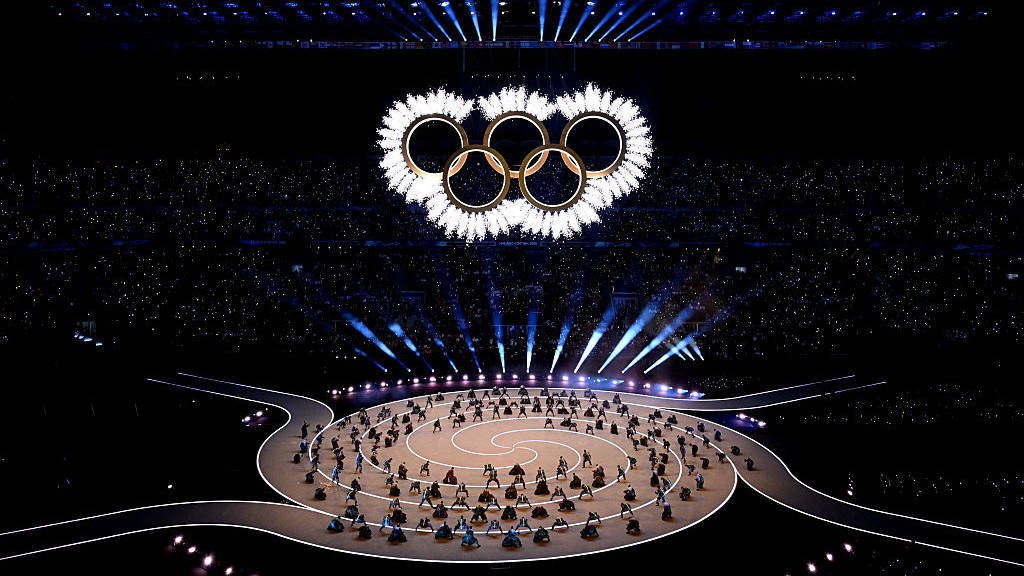 Olympic rings are the top of the image surrounded by fireworks with people below in a spiral formation.