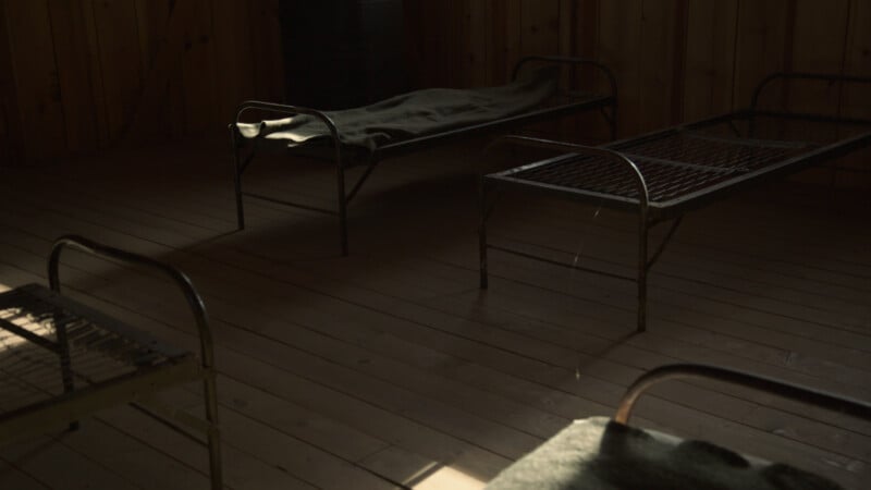 A dimly lit wooden room with three simple metal bed frames, two without mattresses and one with a blanket. Sunlight filters in, casting shadows across the wooden floor.
