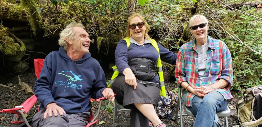 Three people sit in camp chairs outdoors. They are smiling and laughing.