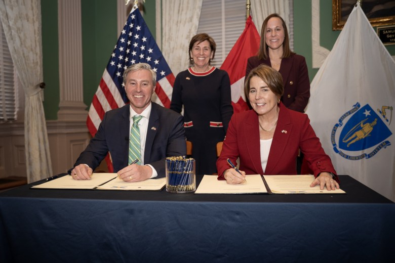 A smiling middle aged white man wearing a dark suit jacket, white shirt, and green and yellow tie sits at a table with a dark cloth beside a middle aged white woman with light brown hair wearing a dark red jacket and white shirt. The pair have papers in front of them, pens poised to sign. Standing behind them are two smiling white women.