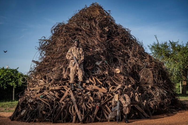 Two soldiers in camouflage uniforms pose with rifles in front of a large pile of rusty scrap metal under a clear blue sky, with trees and greenery visible in the background.