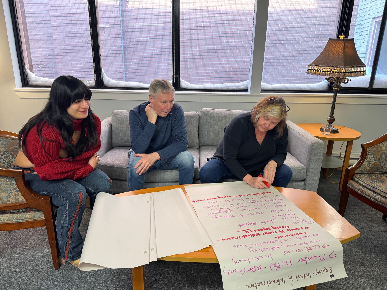 Three people; a man and two women are seated bent over a table with big sheets of paper. One of the women is writing on the paper while the other people look on.