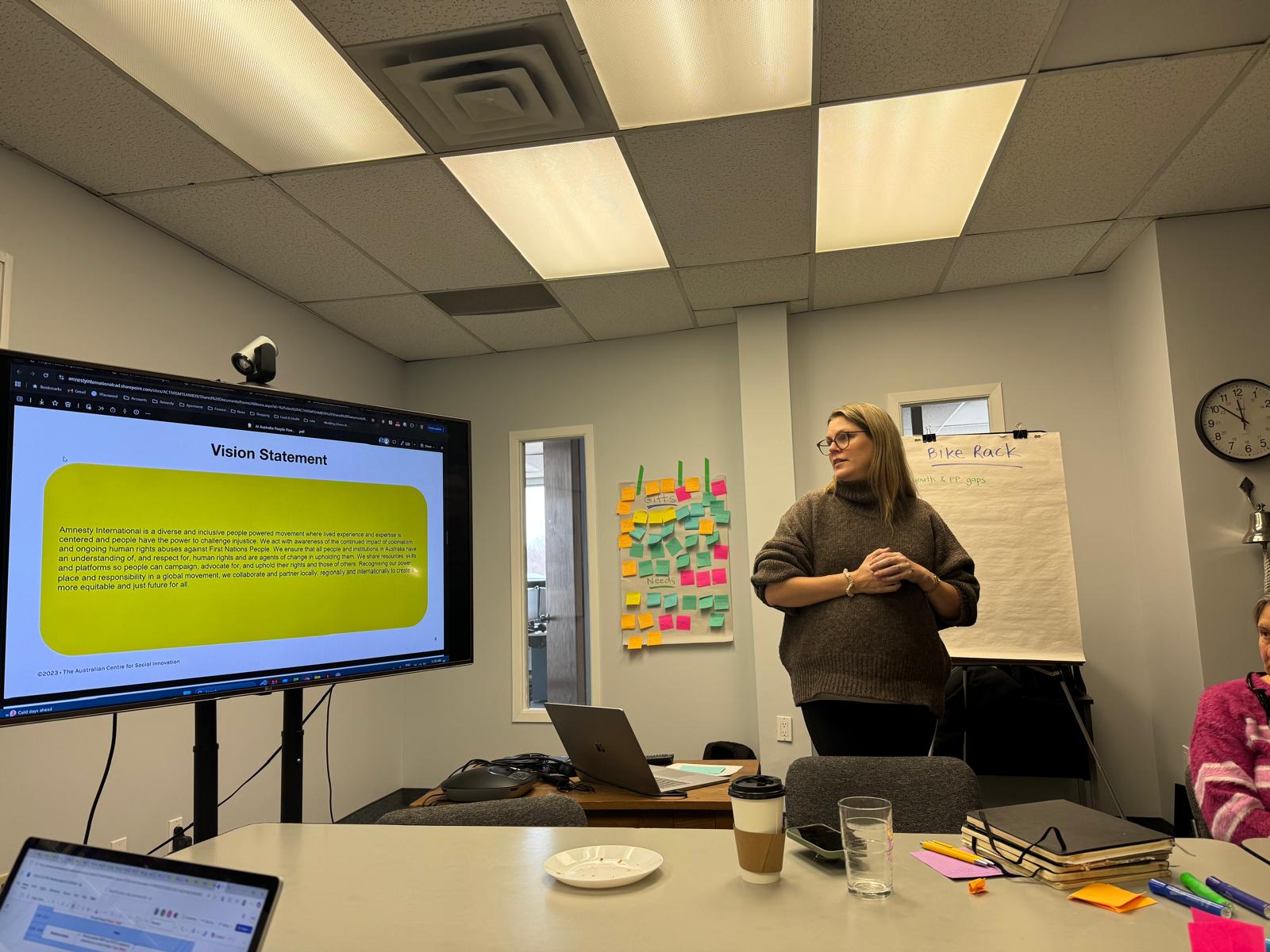 A woman stands in front of a projector screen which has a text block titled Mission Statement