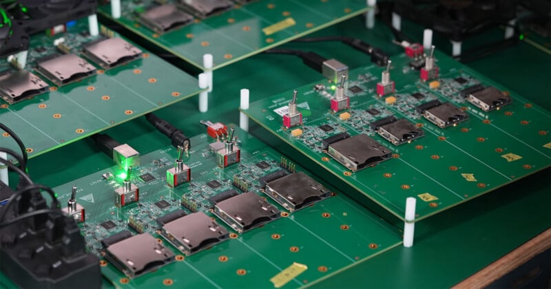 Close-up of green circuit boards with various electronic components, including ports, connectors, chips, and a glowing green LED, arranged on a table for assembly or testing.