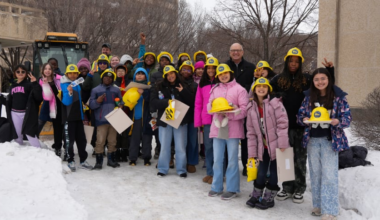 Students in winter coats and yellow hard hats stand outside in the snow with the mayor and a large snow clearing vehicle behind them.