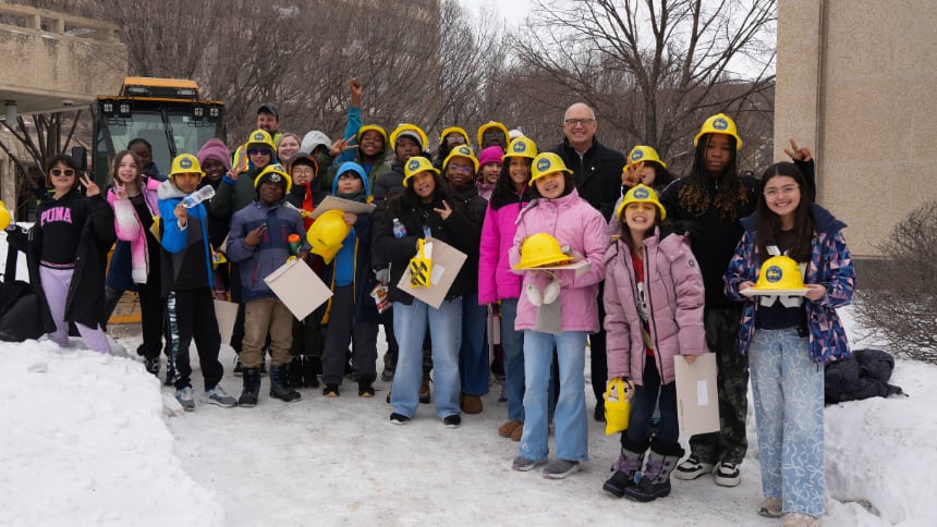 Students in winter coats and yellow hard hats stand outside in the snow with the mayor and a large snow clearing vehicle behind them.