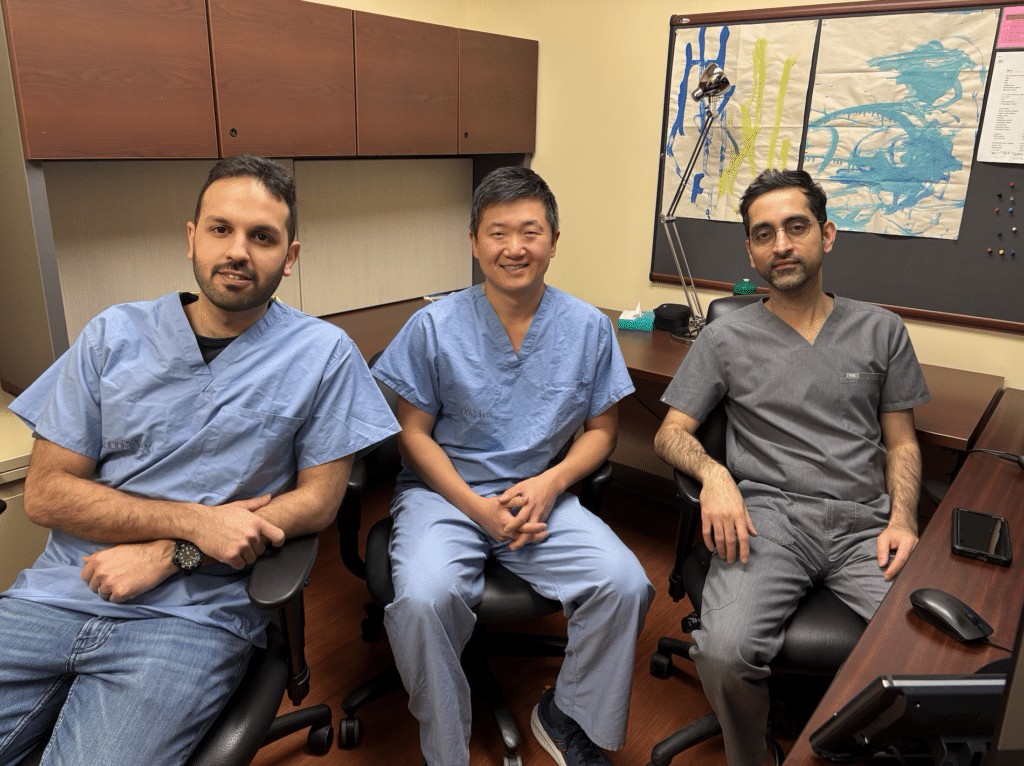 three male doctors wearing blue scrubs sit next to each other in an office.