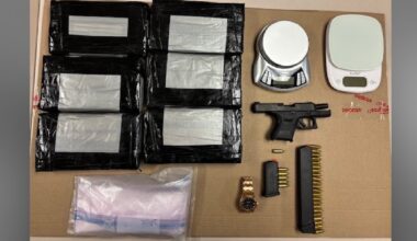 Items  seized by police laid out on a table including several black plastic-wrapped packages, a small handgun, ammunition magazines and loose rounds, a wristwatch, electronic components, a roll of tape or container, and a small digital scale.