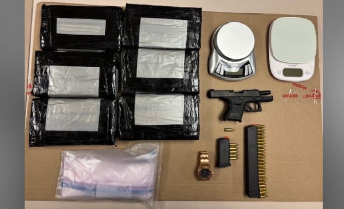 Items  seized by police laid out on a table including several black plastic-wrapped packages, a small handgun, ammunition magazines and loose rounds, a wristwatch, electronic components, a roll of tape or container, and a small digital scale.