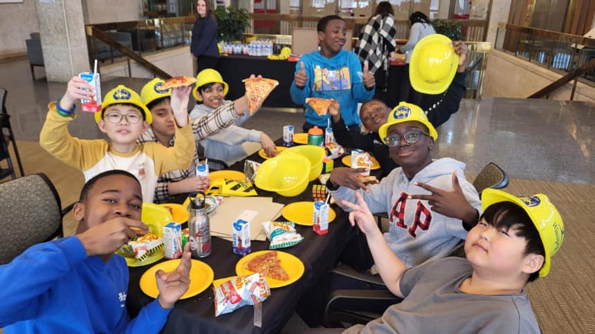 Students wearing yellow hard hats sit around a table posing and eating pizza and drinking juice boxes.