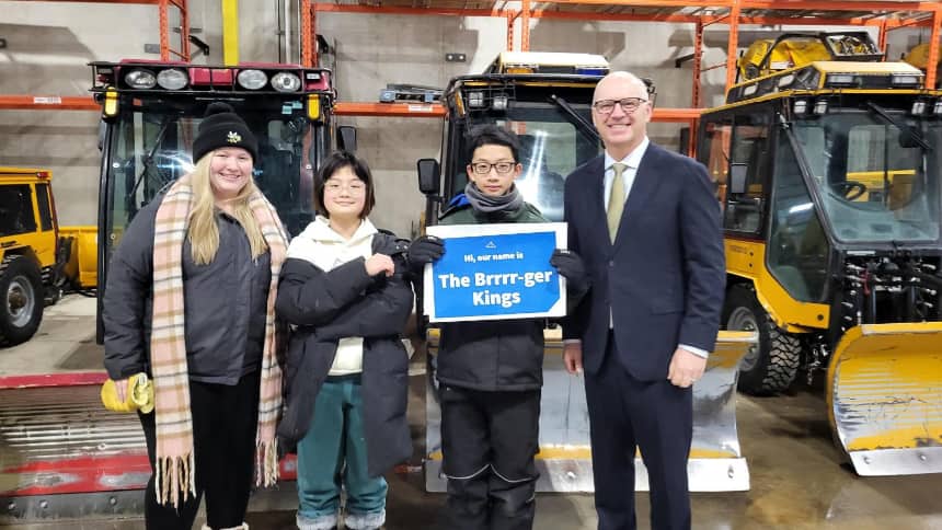 Two students stand in front of snowplows holding a blue sign that says “The Brrrr-ger Kings,” with their teacher and mayor beside them.