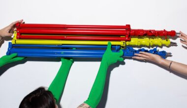 Four hands, two in green gloves, hold three folded metal stands in red, yellow, and blue against a white background.
