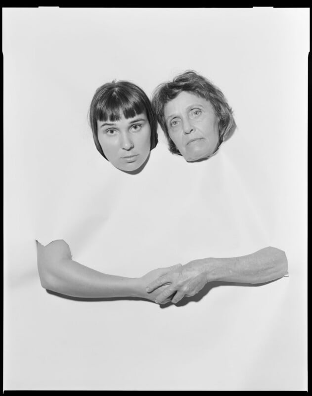 A young woman and an older woman appear to share a single white garment, with only their heads and clasped hands visible against a seamless white background. Their expressions are calm and serious.