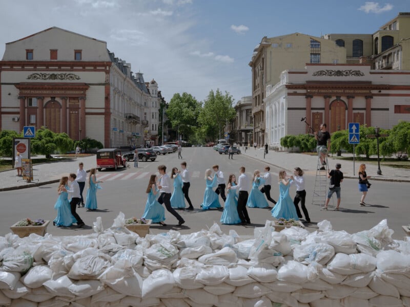 Young couples in blue and white costumes dance in a street flanked by historic buildings, while sandbags line the foreground and a photographer captures the scene from a ladder. The sky is partly cloudy.