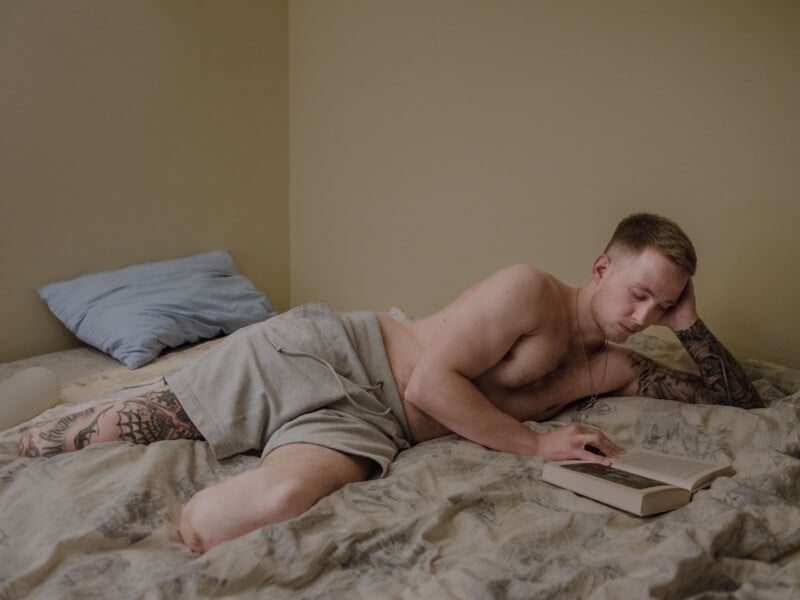 A shirtless man with tattoos reclines on a bed, propped up on one arm, wearing gray shorts and reading a book. The bed has light-colored sheets and a blue pillow, and the room has beige walls.