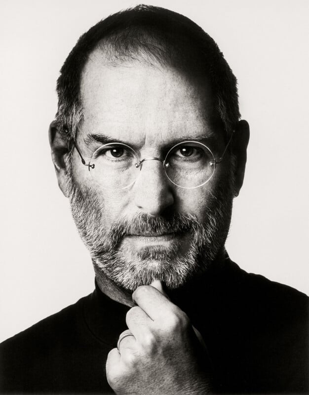 A man with glasses and a beard looks directly at the camera, wearing a dark shirt. He holds his chin with one hand in a thoughtful pose against a plain, light background.