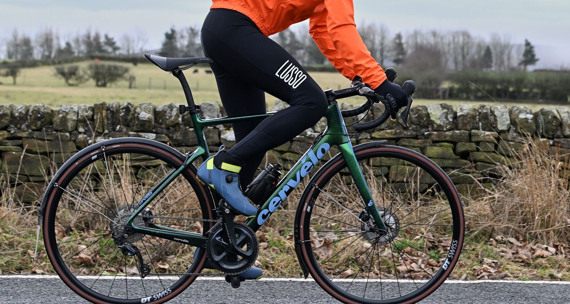 Man wearing black tights, blue shoes and an orange jacket riding a green road bike